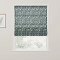 Textile Roman blind with a William Morris-inspired design featuring intertwining light green and white botanical patterns on a dark blue background covering a window in a room, with a gold-framed botanical print visible on the adjacent wall