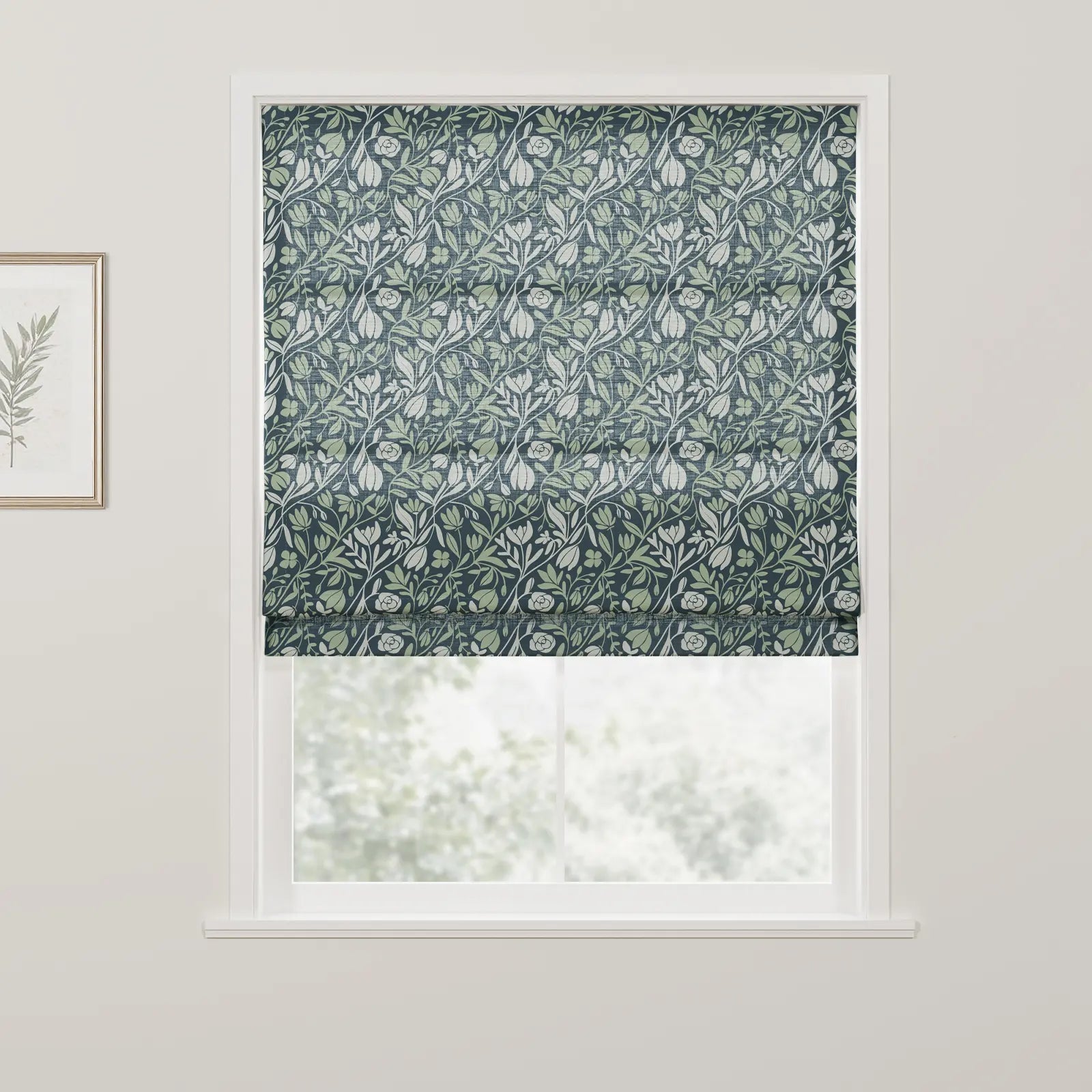 Textile Roman blind with a William Morris-inspired design featuring intertwining light green and white botanical patterns on a dark blue background covering a window in a room, with a gold-framed botanical print visible on the adjacent wall