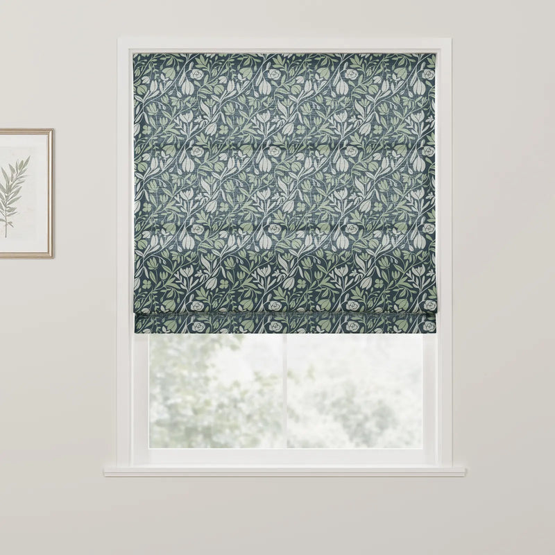 Textile Roman blind with a William Morris-inspired design featuring intertwining light green and white botanical patterns on a dark blue background covering a window in a room, with a gold-framed botanical print visible on the adjacent wall