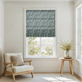 Photograph depicting a bright minimalist room interior with a window dressed in a dark blue-green and sage green William Morris style floral roman blind, complemented by a light cream wooden armchair and a round timber side table displaying a vase with fresh green sprigs