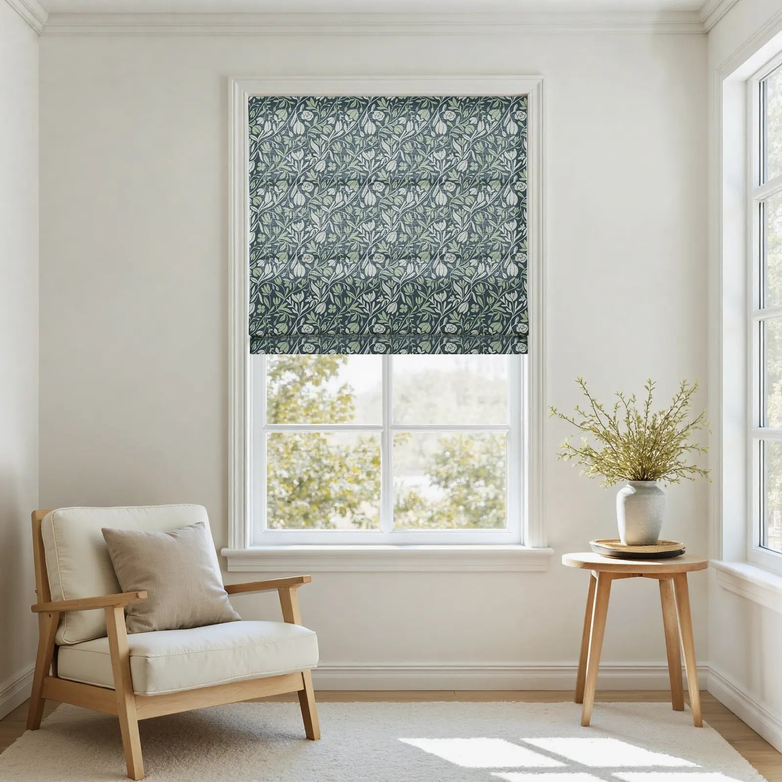 Photograph depicting a bright minimalist room interior with a window dressed in a dark blue-green and sage green William Morris style floral roman blind, complemented by a light cream wooden armchair and a round timber side table displaying a vase with fresh green sprigs