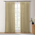 Full-length beige pleated linen curtains on a black rod frame a window overlooking trees with a wooden fluted side table, black vase, and white candle to the right