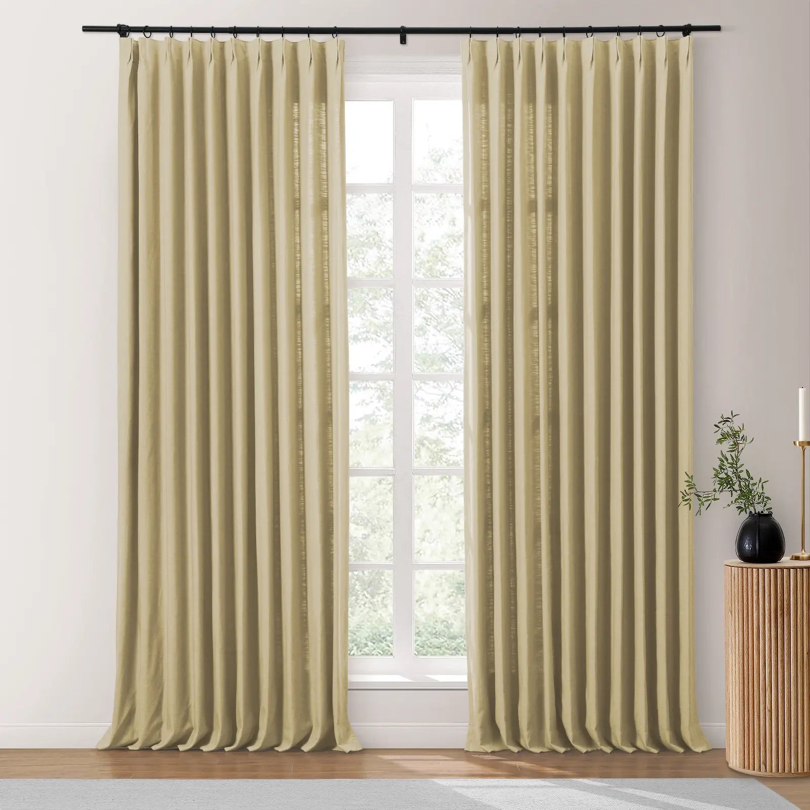 Full-length beige pleated linen curtains on a black rod frame a window overlooking trees with a wooden fluted side table, black vase, and white candle to the right