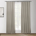 Neutral beige ring-top blackout curtains hanging on a black rod, framing a white paned window looking out to blurred green trees, with an abstract black and white artwork on a white wall and a textured natural fiber rug on a dark wood floor