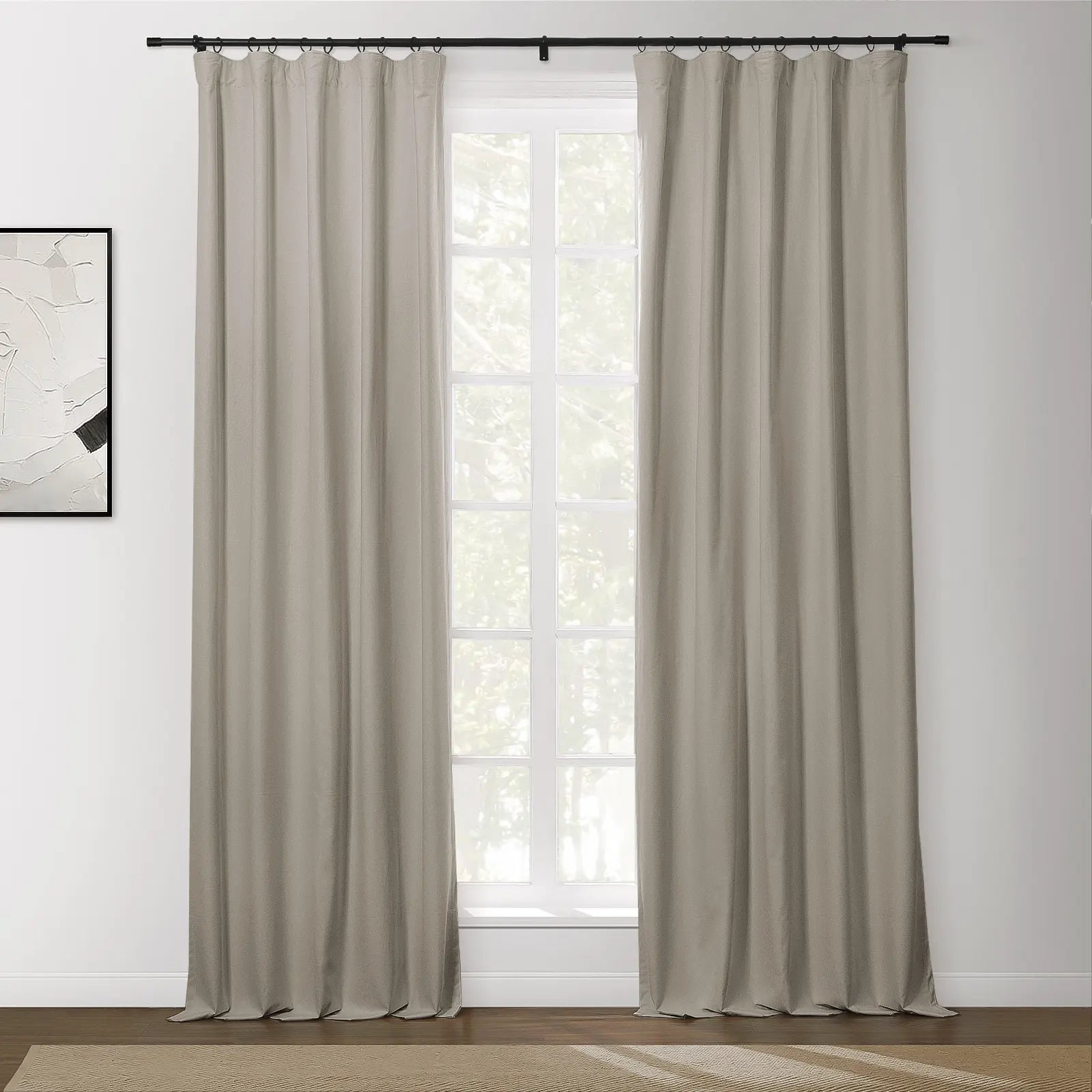 Neutral beige ring-top blackout curtains hanging on a black rod, framing a white paned window looking out to blurred green trees, with an abstract black and white artwork on a white wall and a textured natural fiber rug on a dark wood floor
