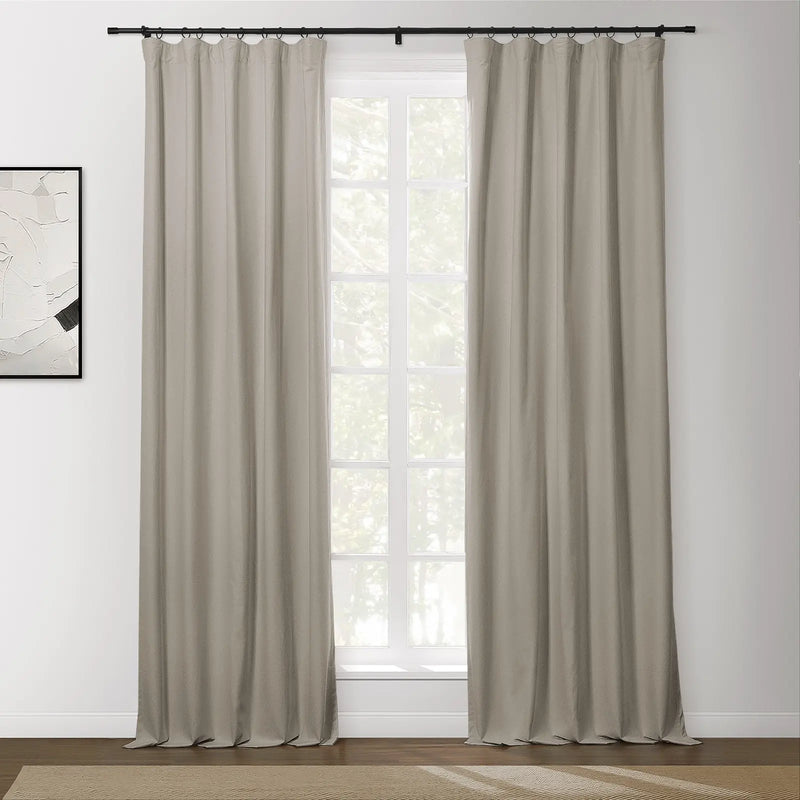 Neutral beige ring-top blackout curtains hanging on a black rod, framing a white paned window looking out to blurred green trees, with an abstract black and white artwork on a white wall and a textured natural fiber rug on a dark wood floor