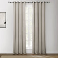 A pair of light beige grommet top curtains on a black rod partially cover a window with white grilles, revealing greenery outside, next to a modern abstract painting on a white wall above a dark wood floor with a woven rug