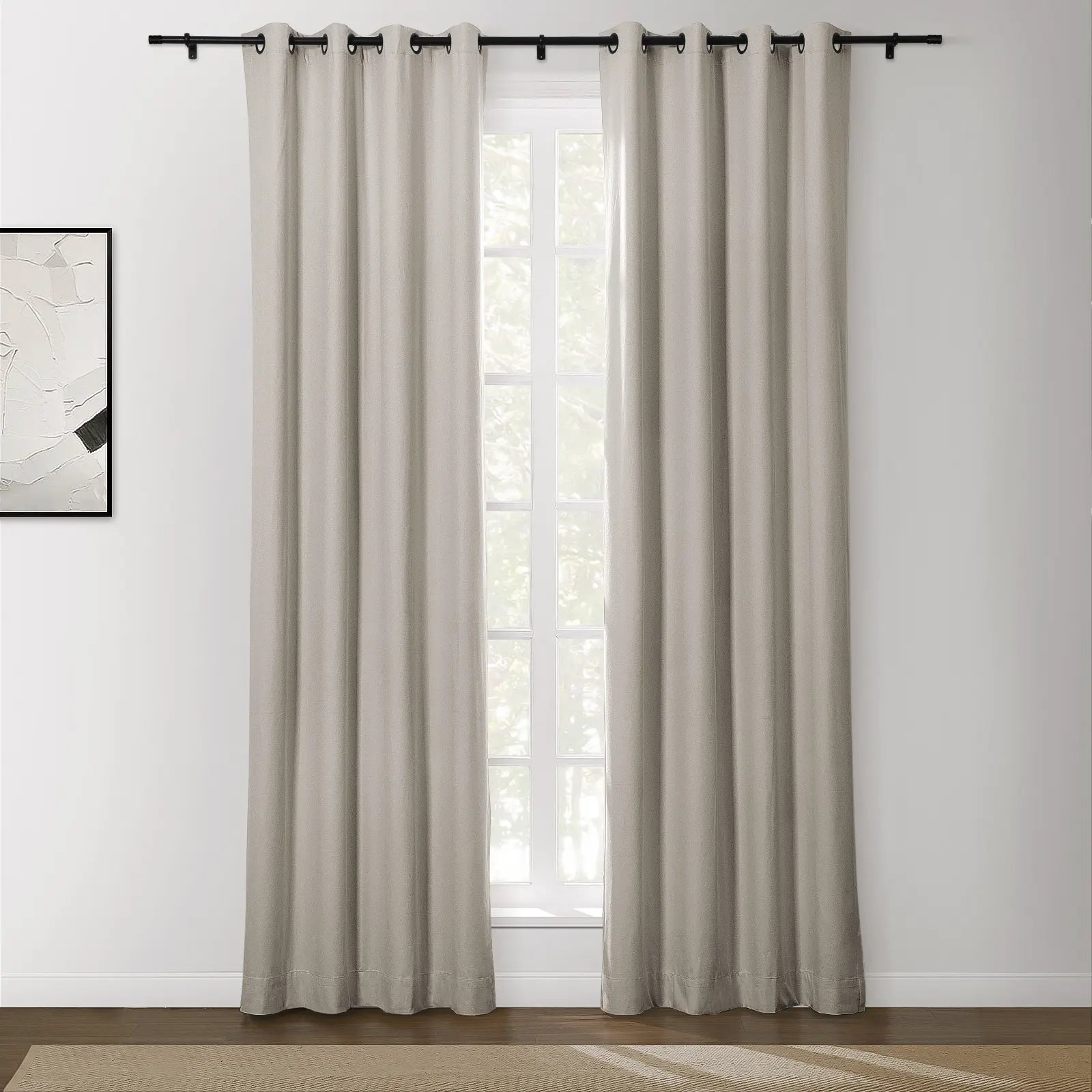 A pair of light beige grommet top curtains on a black rod partially cover a window with white grilles, revealing greenery outside, next to a modern abstract painting on a white wall above a dark wood floor with a woven rug