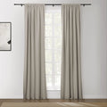 A pair of light beige rod pocket curtains on a black rod frame a sunlit multi-pane window revealing lush green trees, with a subtle white and black abstract painting on the left wall and a textured beige rug on the hardwood floor