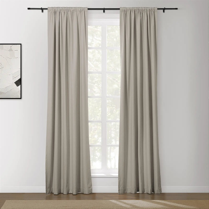 A pair of light beige rod pocket curtains on a black rod frame a sunlit multi-pane window revealing lush green trees, with a subtle white and black abstract painting on the left wall and a textured beige rug on the hardwood floor