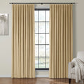 Beige pleated velvet curtains on a black rod frame a large window in a modern living room featuring abstract tree wall art and a gray armchair with a white throw