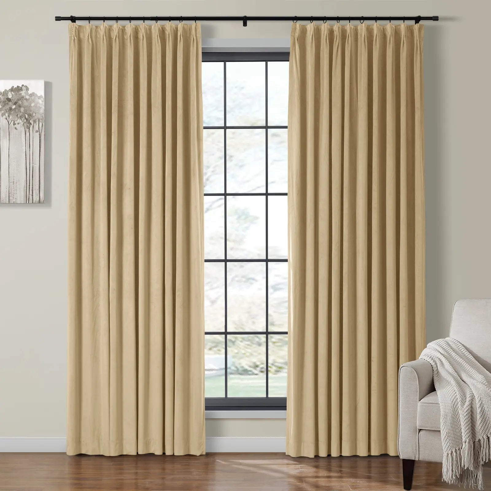 Beige pleated velvet curtains on a black rod frame a large window in a modern living room featuring abstract tree wall art and a gray armchair with a white throw