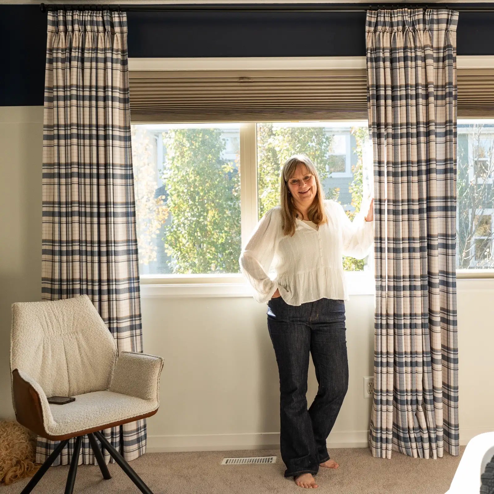 Smiling woman stands barefoot by a sunlit window with blue and beige plaid curtains, a cream bouclé armchair, and a dark blue accent wall in a modern living space