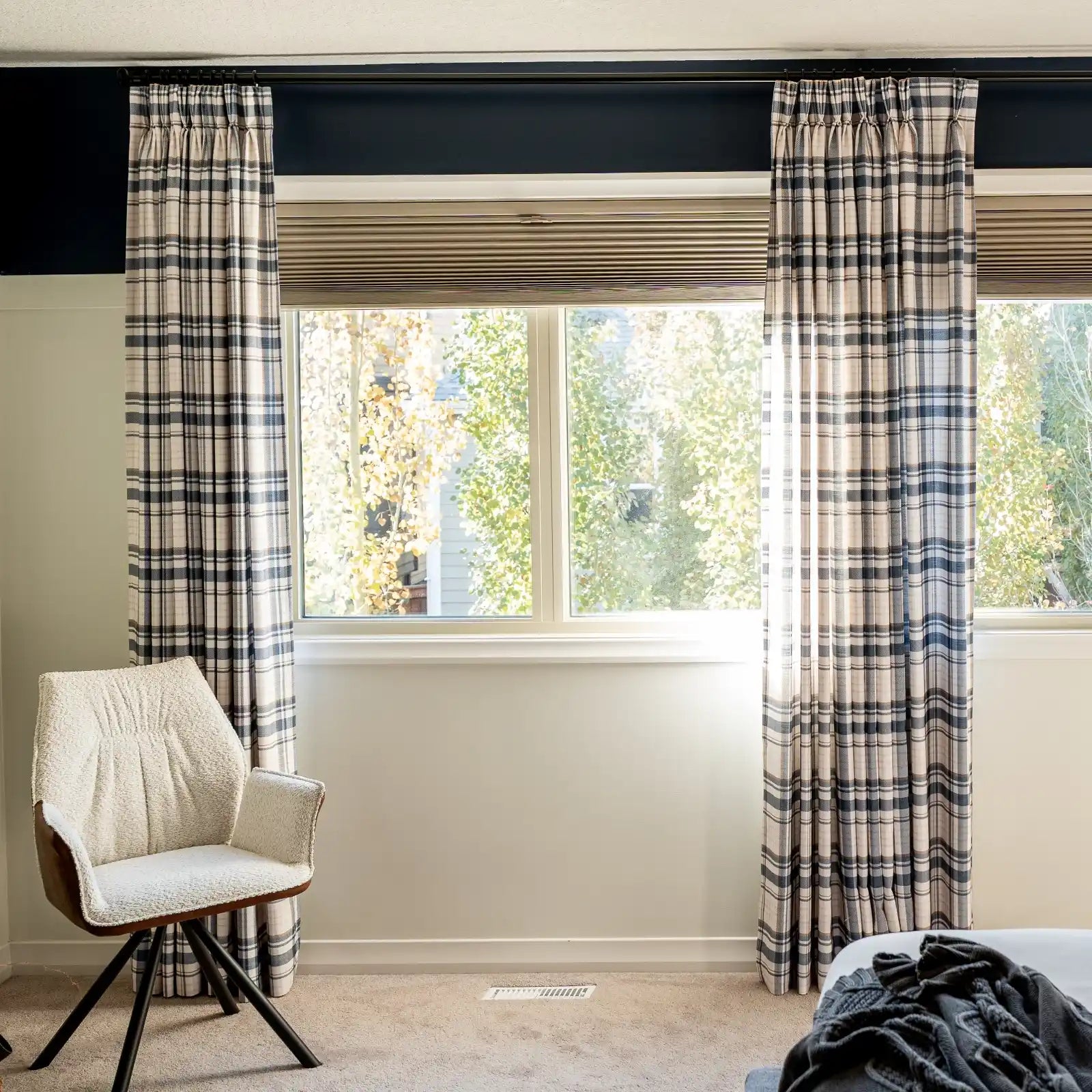 A cozy bedroom interior with a large window displaying fall trees outside, featuring a dark blue accent wall above pleated shades, black and white plaid curtains, and a white boucle upholstered accent chair with a dark splayed base