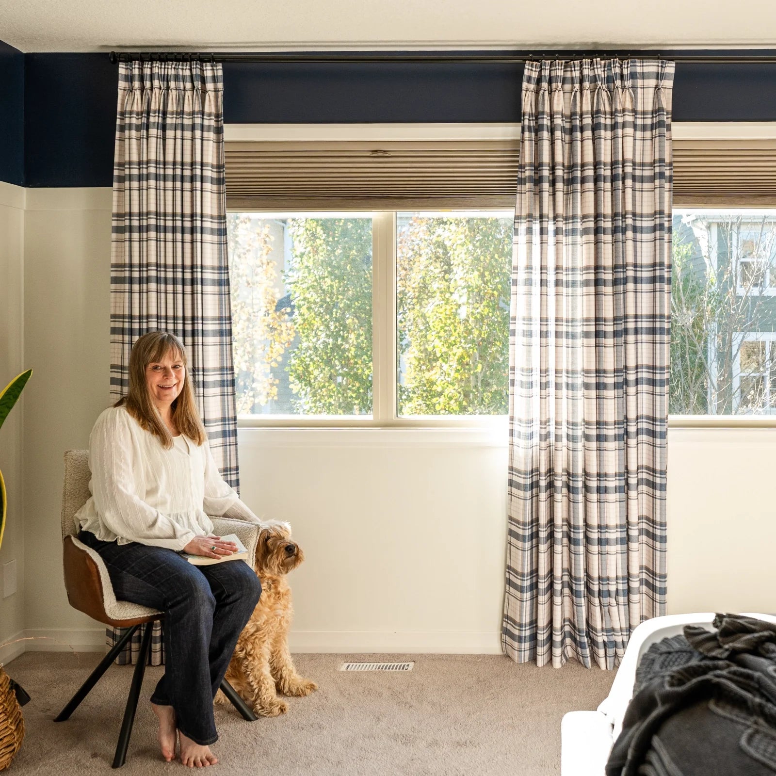 Photograph of a smiling woman and her fluffy light brown dog in a cozy room by a large window with navy and beige plaid curtains, featuring a distinct dark navy and cream two-tone wall