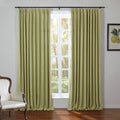 A room interior featuring floor-length olive green pinch-pleat curtains framing a window with a sunlit view of green trees, alongside a framed botanical lemon print