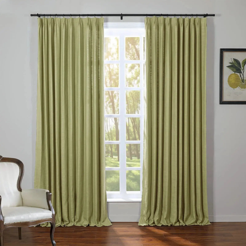 A room interior featuring floor-length olive green pinch-pleat curtains framing a window with a sunlit view of green trees, alongside a framed botanical lemon print