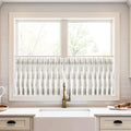 White cafe curtains with a grey leaf pattern on a brass rod over a white farmhouse sink and brass faucet in a brightly lit kitchen with subway tile backsplash