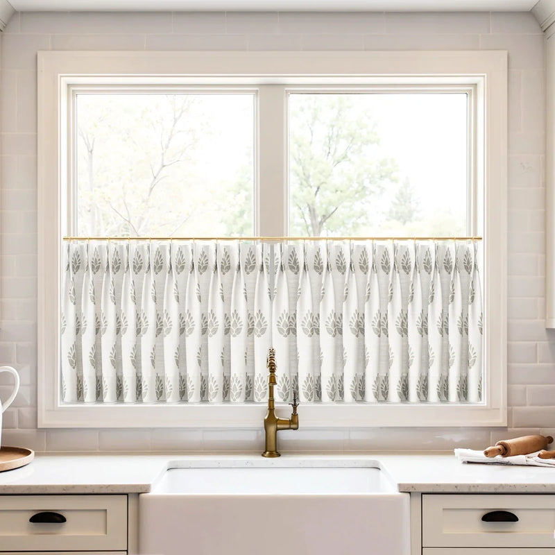 White cafe curtains with a grey leaf pattern on a brass rod over a white farmhouse sink and brass faucet in a brightly lit kitchen with subway tile backsplash