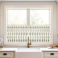 Photograph of a modern kitchen window featuring a white cafe curtain with an olive green leaf pattern, centered above a white farmhouse sink and a brushed brass gooseneck faucet