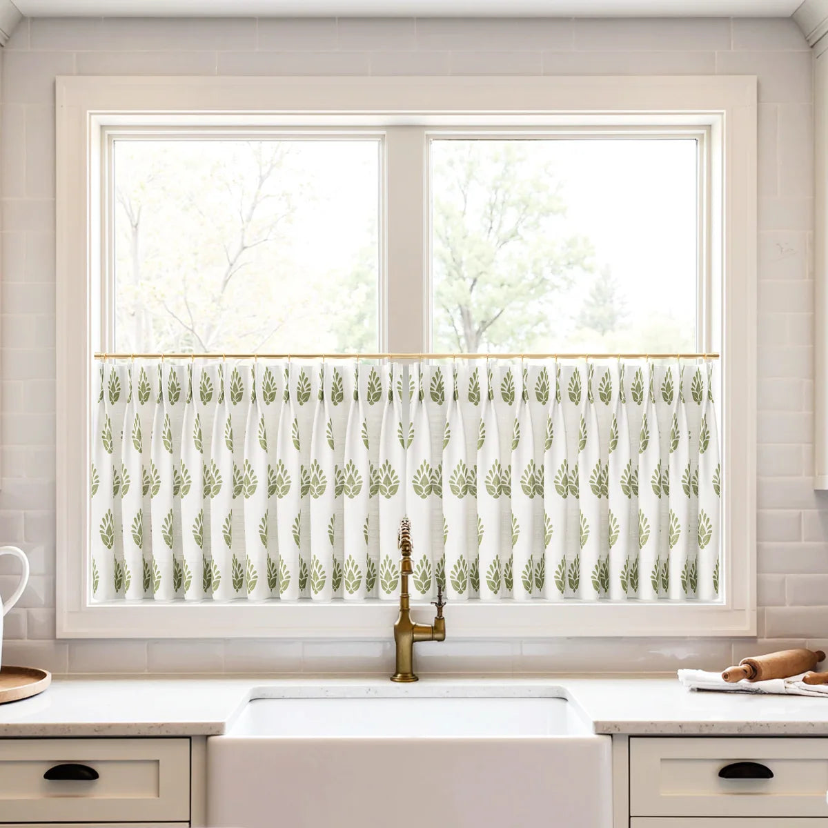 Photograph of a modern kitchen window featuring a white cafe curtain with an olive green leaf pattern, centered above a white farmhouse sink and a brushed brass gooseneck faucet