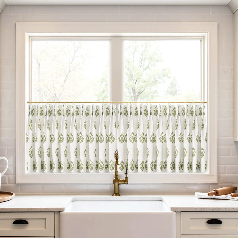 Photograph of a modern kitchen window featuring a white cafe curtain with an olive green leaf pattern, centered above a white farmhouse sink and a brushed brass gooseneck faucet