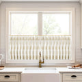 A modern kitchen window above a white farmhouse sink featuring white cafe curtains with a gold leaf pattern, a decorative brass faucet, and a white subway tile backsplash