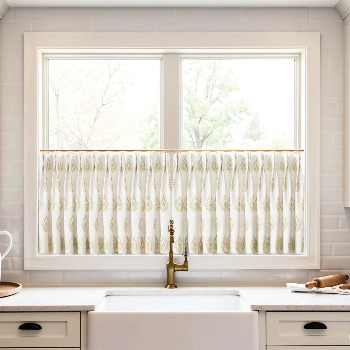A modern kitchen window above a white farmhouse sink featuring white cafe curtains with a gold leaf pattern, a decorative brass faucet, and a white subway tile backsplash