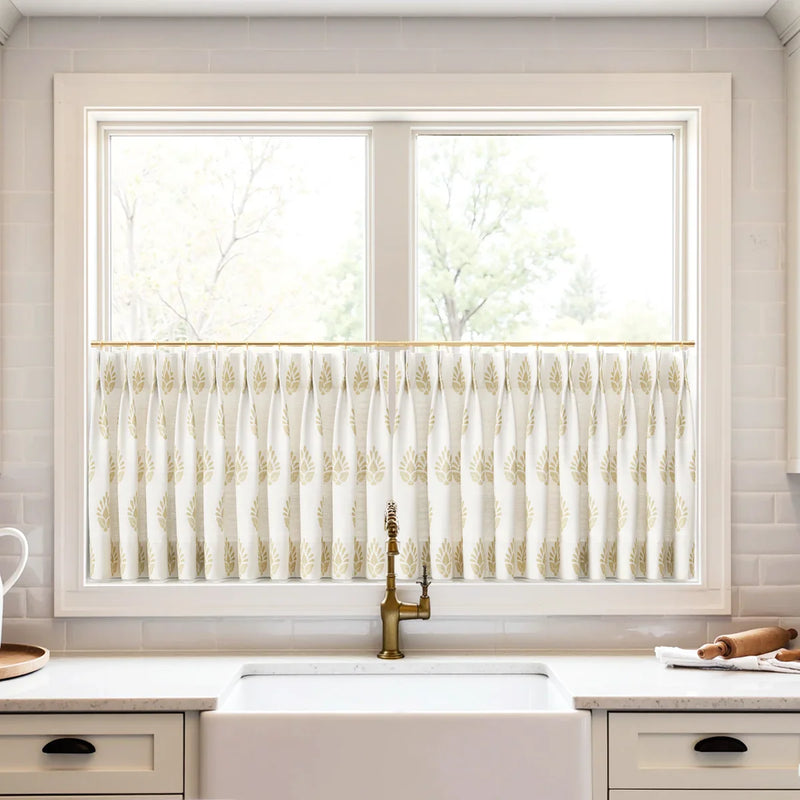 A modern kitchen window above a white farmhouse sink featuring white cafe curtains with a gold leaf pattern, a decorative brass faucet, and a white subway tile backsplash