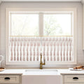 A bright kitchen window with white cafe curtains featuring a reddish-pink leaf pattern on a brass rod, above a white farmhouse sink and a vintage brass faucet