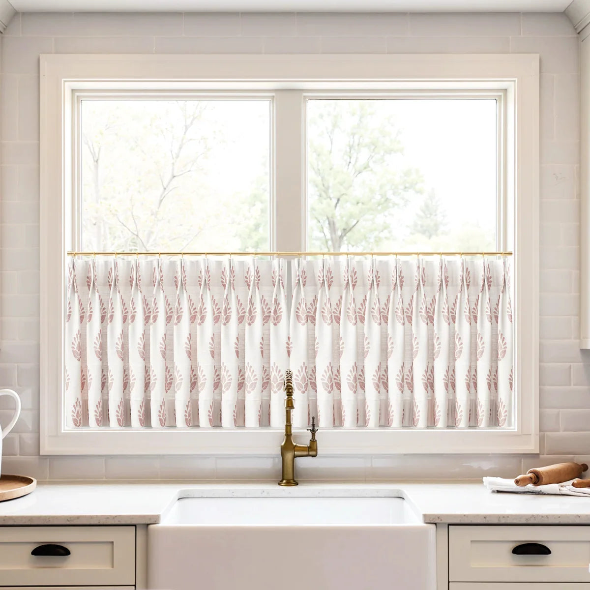A bright kitchen window with white cafe curtains featuring a reddish-pink leaf pattern on a brass rod, above a white farmhouse sink and a vintage brass faucet