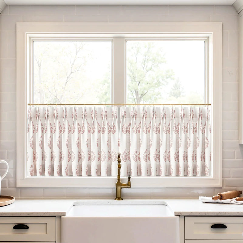 A bright kitchen window with white cafe curtains featuring a reddish-pink leaf pattern on a brass rod, above a white farmhouse sink and a vintage brass faucet