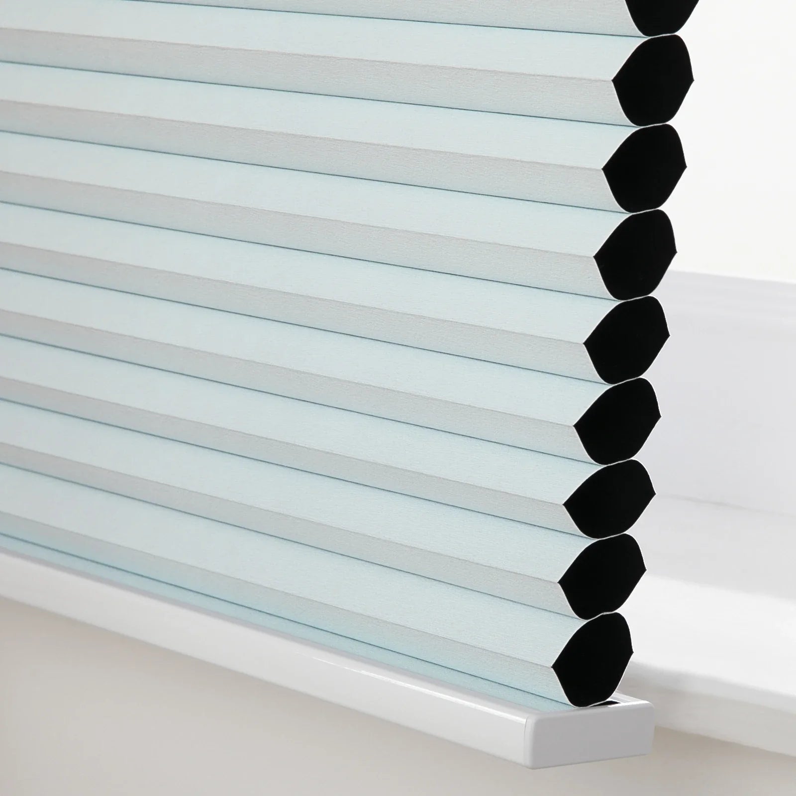 Close-up photograph of a light blue cellular window Blind featuring visible black honeycomb cells resting on a white windowsill