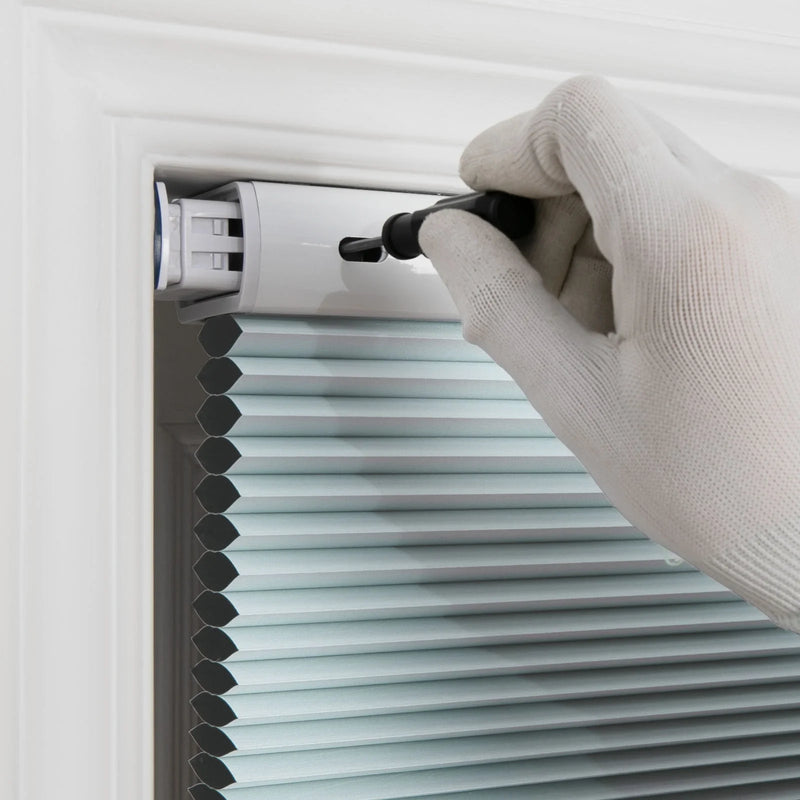 A white gloved hand uses a black adjustment tool on a light blue pleated cellular window Blind installed in a white frame