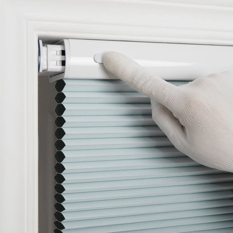 Photography of a white gloved hand pressing a white button on the top rail of light blue cellular Blinds featuring visible hexagonal pleats within a white window frame