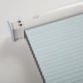 Product photography of a modern light blue pleated window Blind featuring a white plastic casing with an oval button and a translucent blue end cap on a neutral gray studio background