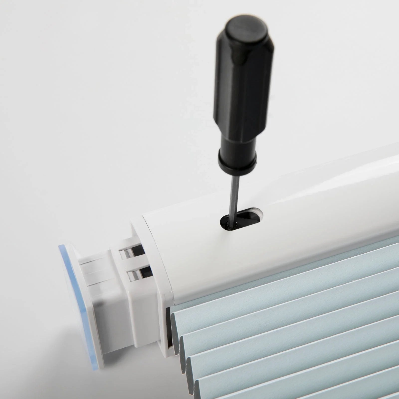 A black screwdriver inserted into a white plastic window Blind headrail mechanism, adjusting a screw above light blue pleated fabric on a bright white surface