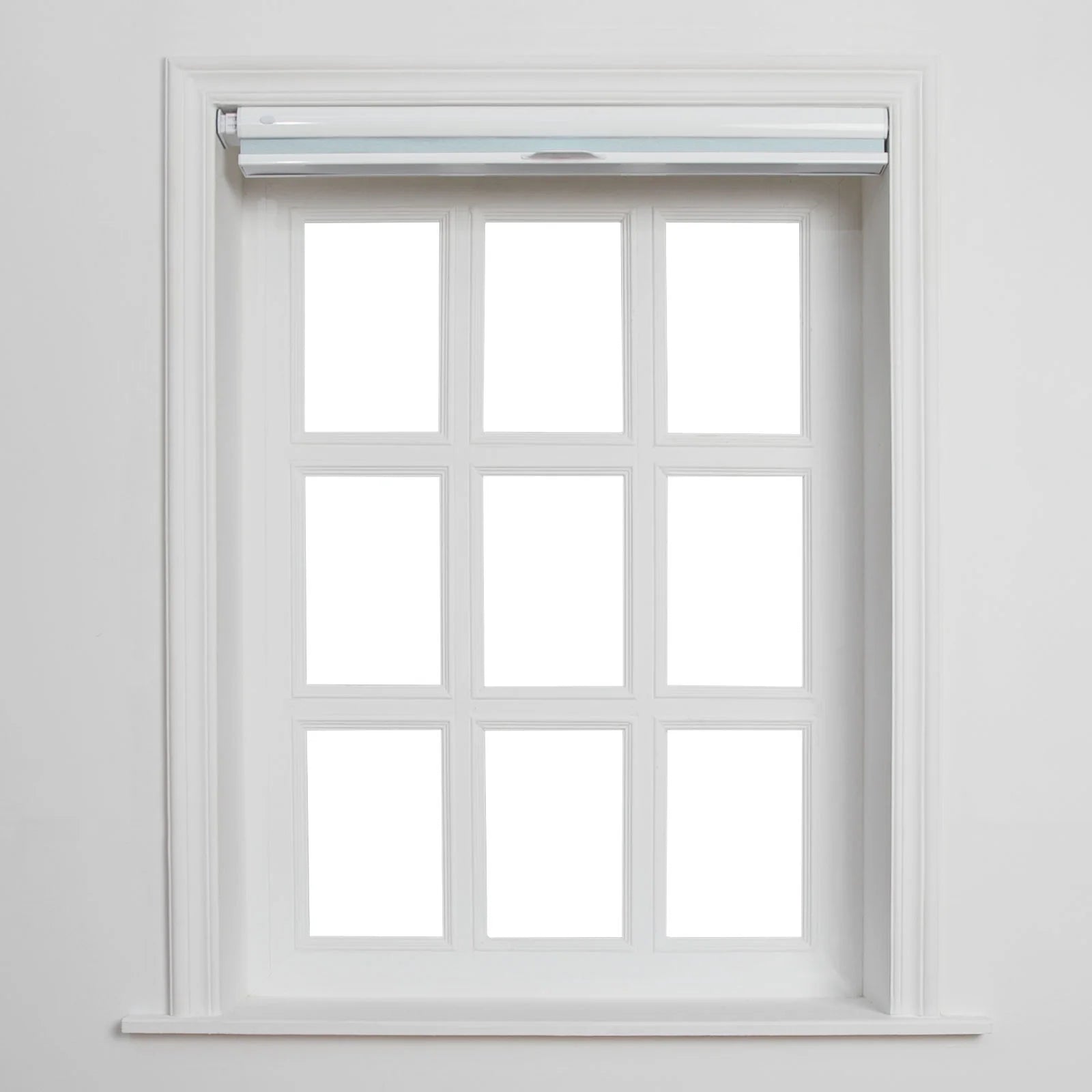 A white-framed window with nine blank panes, featuring a partially rolled-up light blue-grey Blind, and intricate white wooden trim against a clean wall