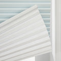 White cellular pleated window Blind partially lowered against a window frame, revealing a light blue pleated Blind behind it, with visible textured fabric and a white top rail