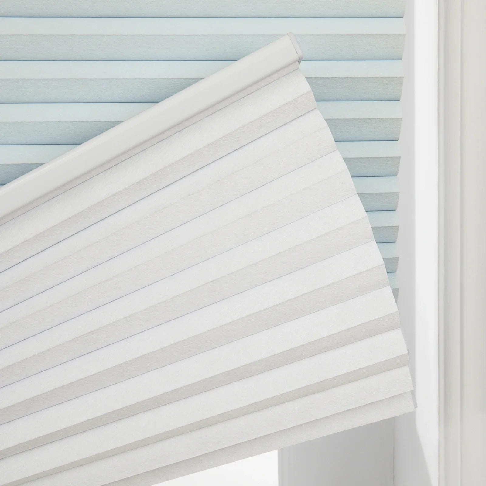 White cellular pleated window Blind partially lowered against a window frame, revealing a light blue pleated Blind behind it, with visible textured fabric and a white top rail