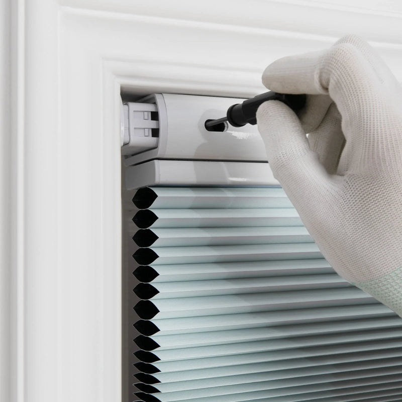 A hand in a white textured glove with green trim uses a black tool to adjust light blue-grey honeycomb cellular Blinds installed in a white window frame