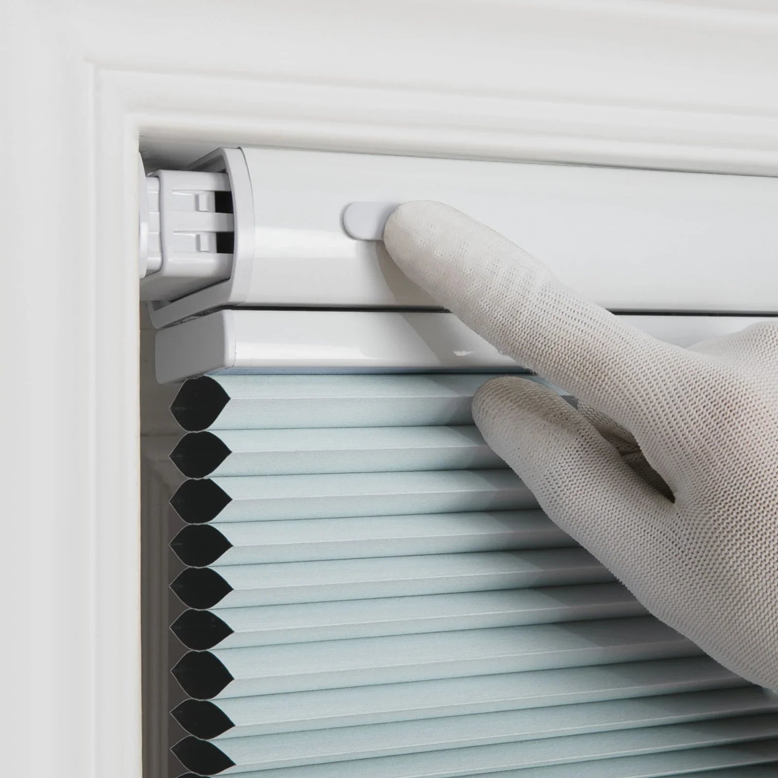 A white gloved hand pressing a control button on the white headrail of light blue cellular honeycomb Blinds installed within a white window frame