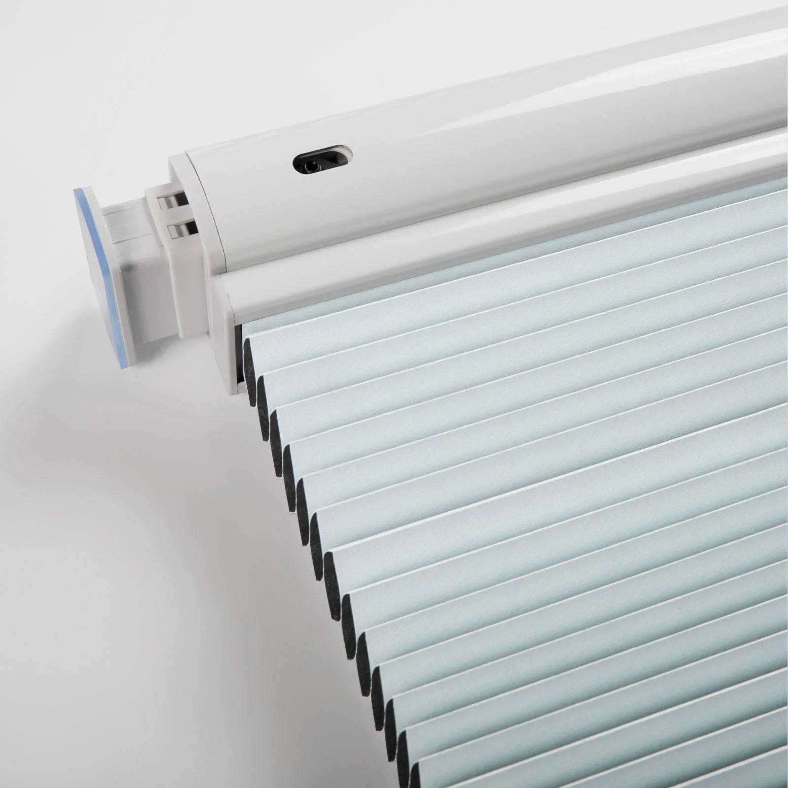 Close-up of a modern white motorized pleated window Blind system with light blue fabric pleats a black sensor on the headrail and a translucent blue end cap