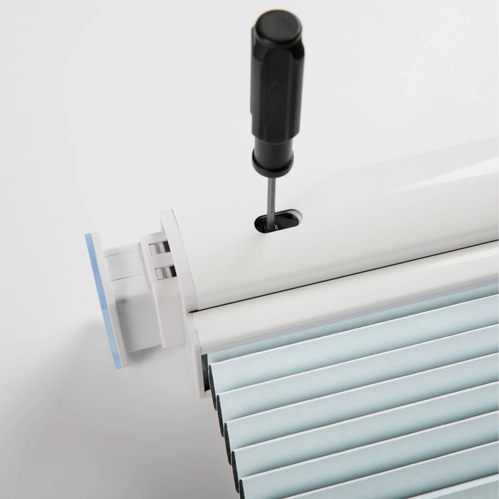 A black-handled screwdriver adjusts a white headrail of a light blue pleated Blind featuring a small blue plastic end tab