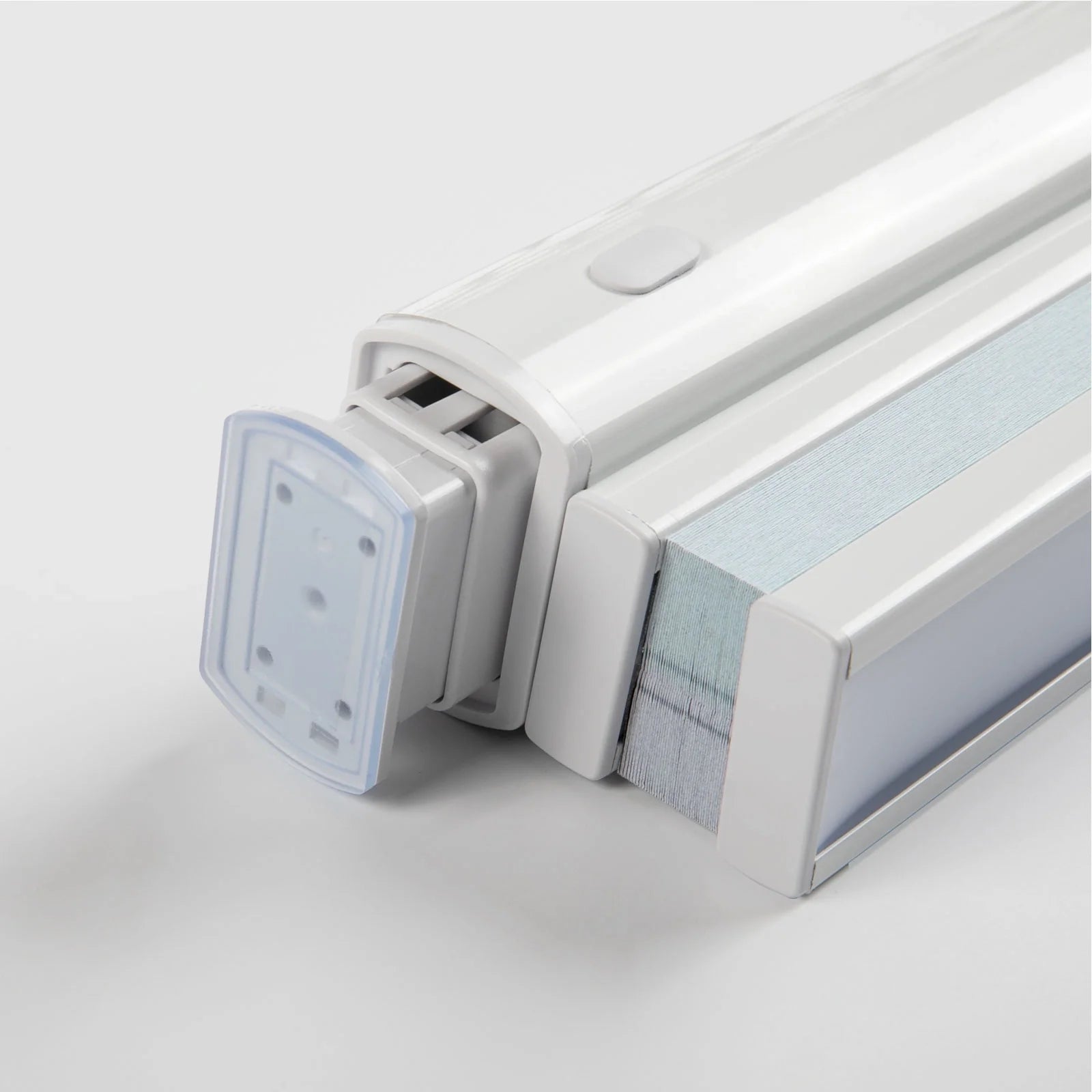 Close-up of a white modern window Blind headrail featuring a clear plastic end cap with mounting holes, a small gray control button on its top surface, and compressed light blue and gray pleated fabric