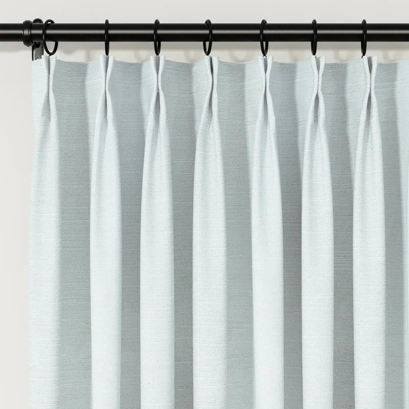 Photograph of light blue-grey textured pinch pleat curtains on a black rod with rings