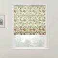 A Roman blind with a light cream background and an intricate floral pattern of rust orange roses yellow flowers and grey-green leaves hangs in a white-framed window overlooking blurred green foliage