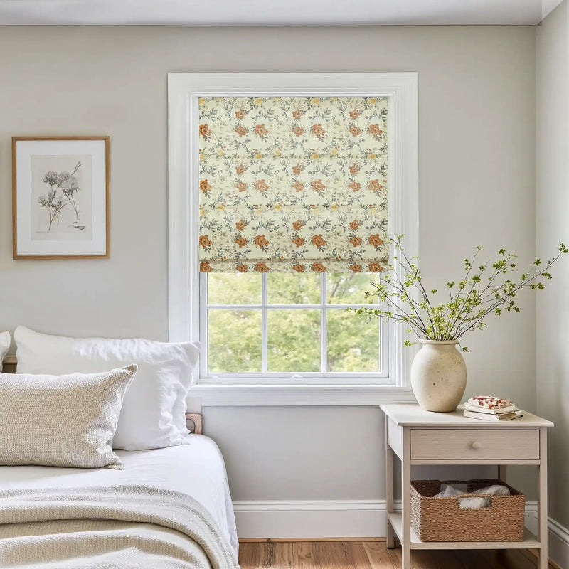 A bright bedroom interior featuring a window with a cream floral Roman shade showcasing orange roses, a white bed with neutral pillows, and a light wooden nightstand holding a large vase of green branches.