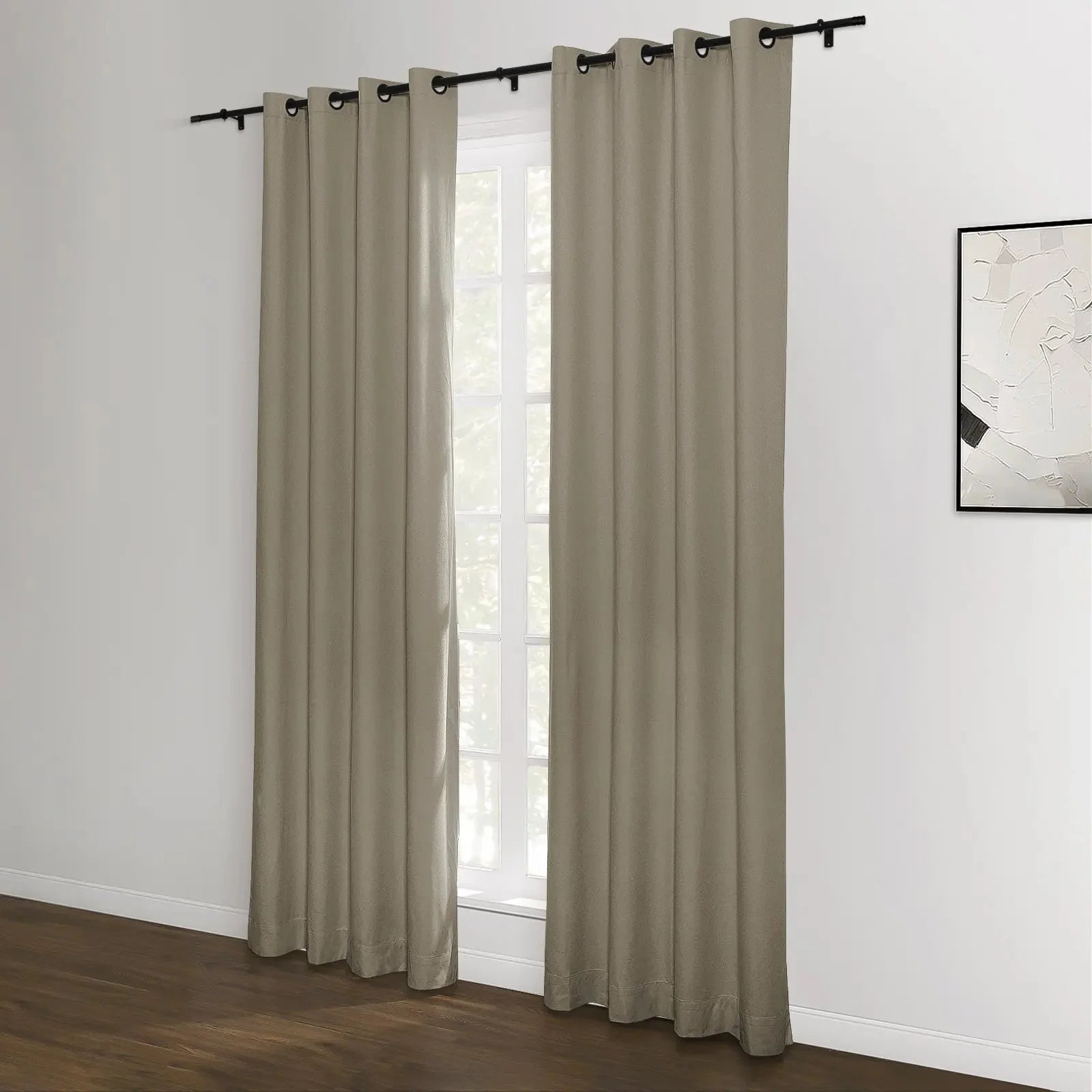 A pair of neutral beige grommet top blackout curtains hang on a black rod in a modern room with a white wall, dark wood floor, and an abstract black and white painting visible