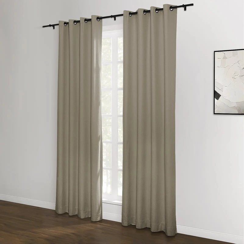 A pair of neutral beige grommet top blackout curtains hang on a black rod in a modern room with a white wall, dark wood floor, and an abstract black and white painting visible